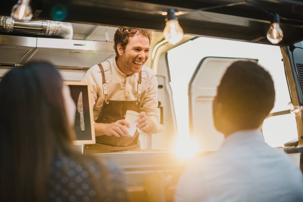 Delighted man with takeaway cups smiling and talking with diverse couple while working in food truck in evening on street