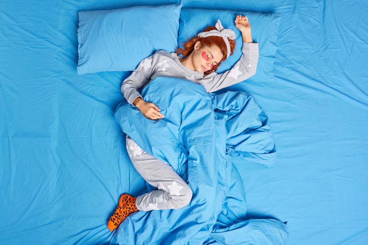 Above view of relaxed redhead woman dressed in pajama headband applied beauty pattches under eyes before sleep to reduce puffiness poses on comfortabled bed feels lazy at home. Peaceful atmosphere