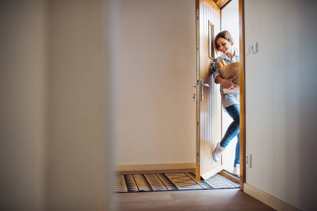 A young woman with groceries in paper shopping bag walking in through front door.