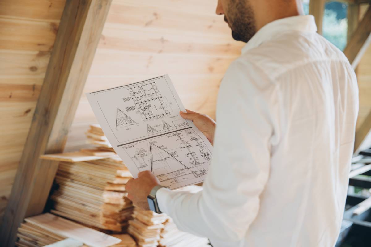 Architect wooden buildings. Man stands under frame wooden house. Man architect studies documentation or blueprints. Guy in protective helmet at construction site. Architect under timber framed house