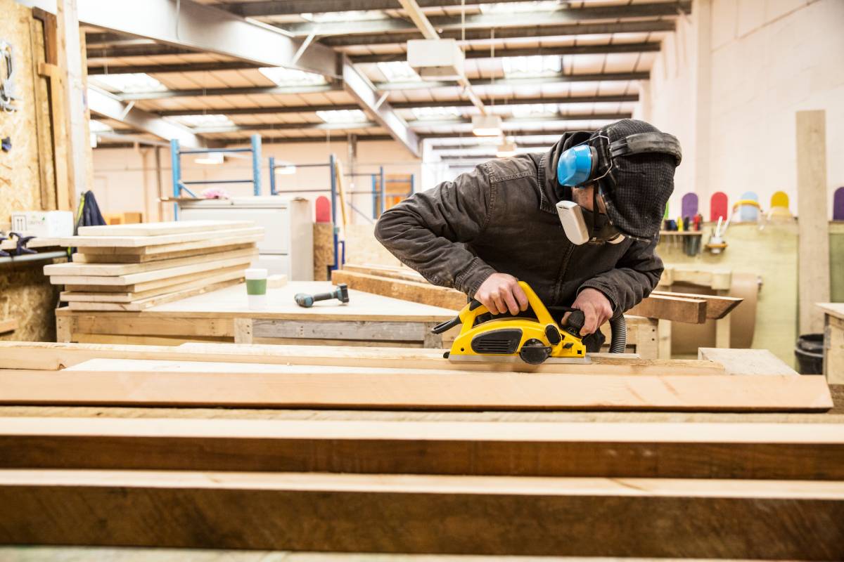 Man wearing ear protectors, protective goggles and dust mask standing in a warehouse, sanding planks of recycled wood. commercial carpentry