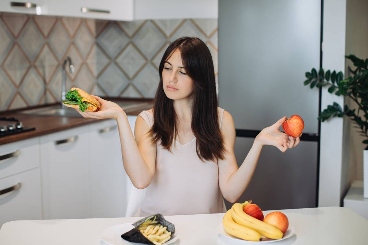 A young woman is choosing between an apple and a burger while sitting at the kitchen table.