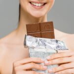 cropped view of smiling young woman holding chocolate bar isolated on grey