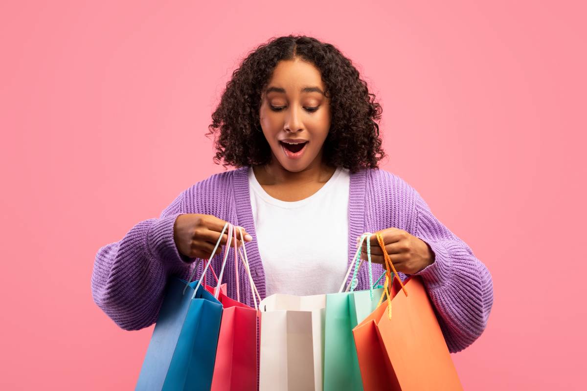 Excited young black woman looking inside shopping bags on pink studio background
