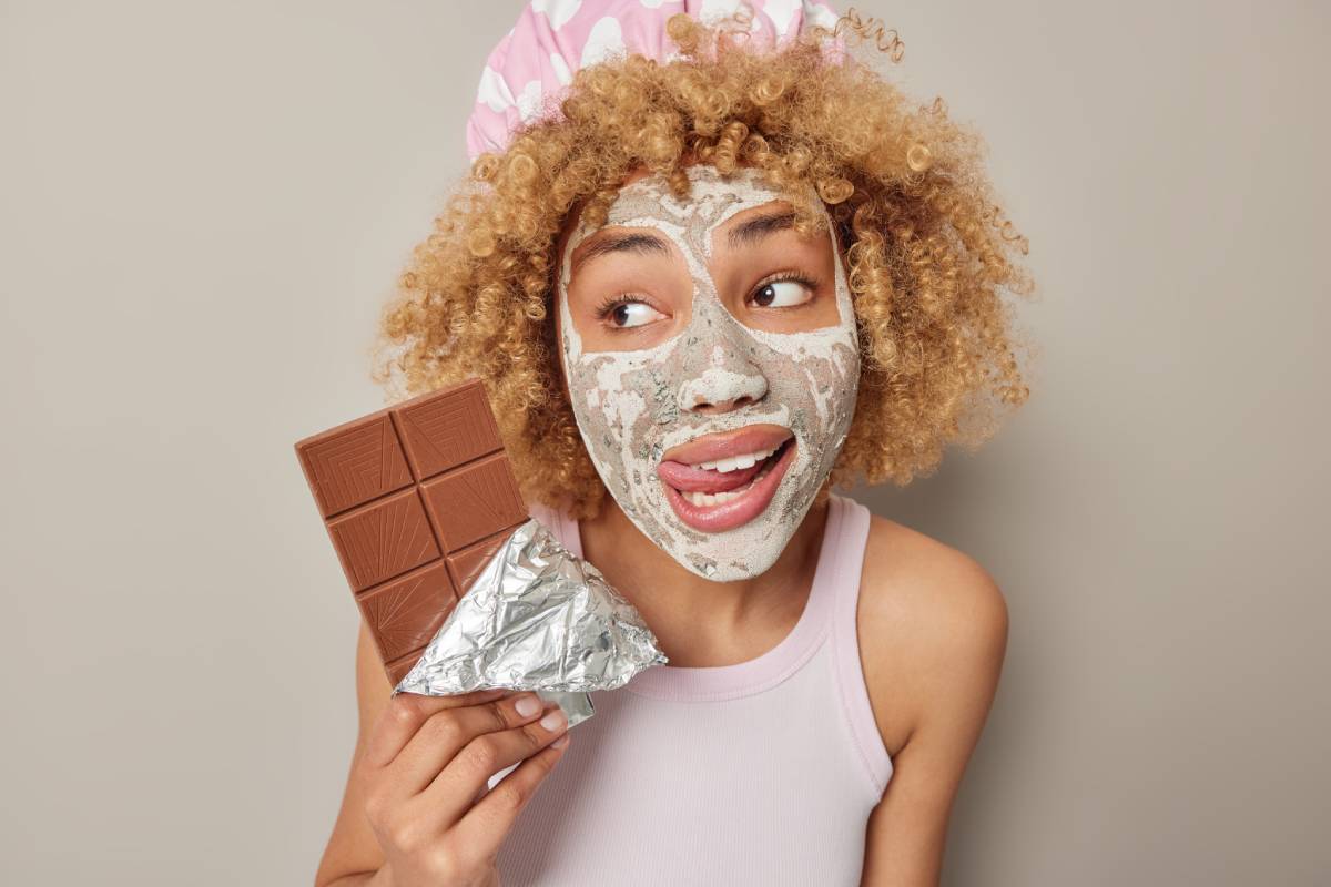 Horizontal shot of curly haired woman bites lips with tongue looks away applies beauty facial mask holds bar of chocolate wears bathhat and t shirt isolated over grey background. Skin care concept