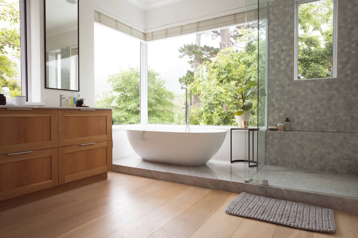 Modern bathroom interior showing free standing bath with large windows showing green trees and plants outside, with white bathtub and silver water tap, grey shower and a table with a plant.